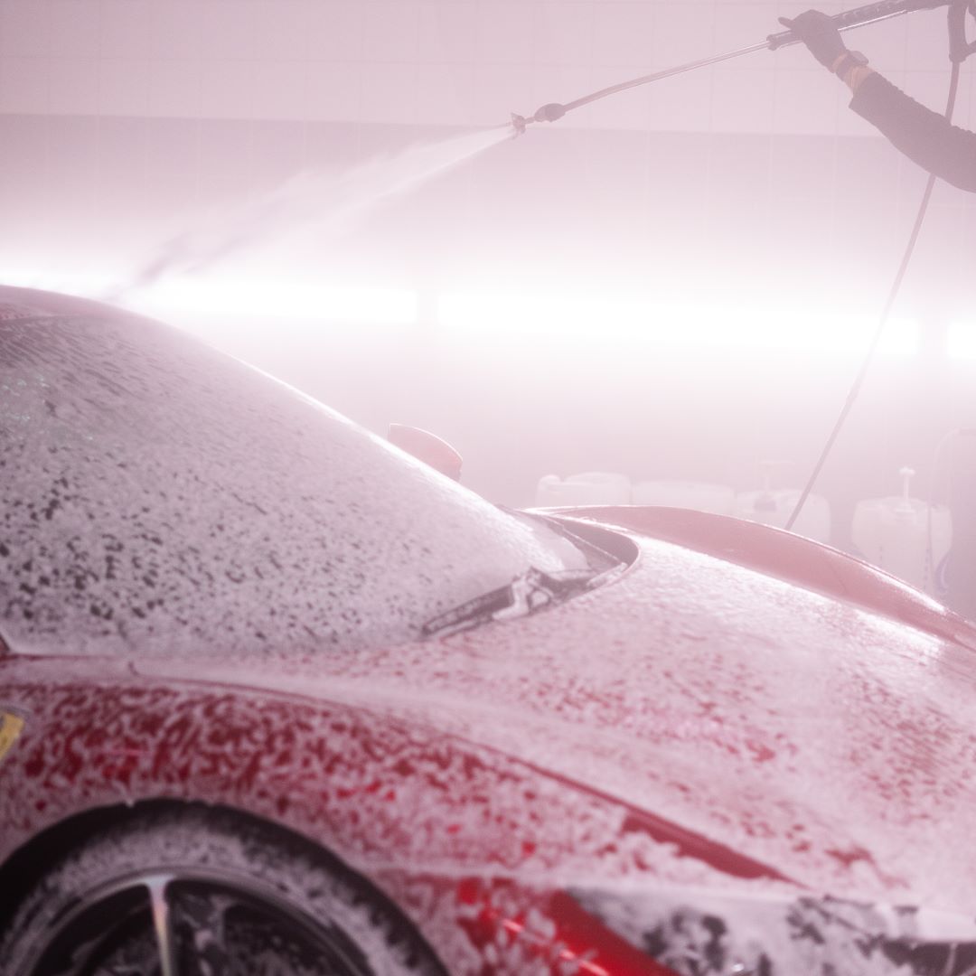 A red sports car being covered with a white soapy cleaning foam by someone using a jetwashing tool