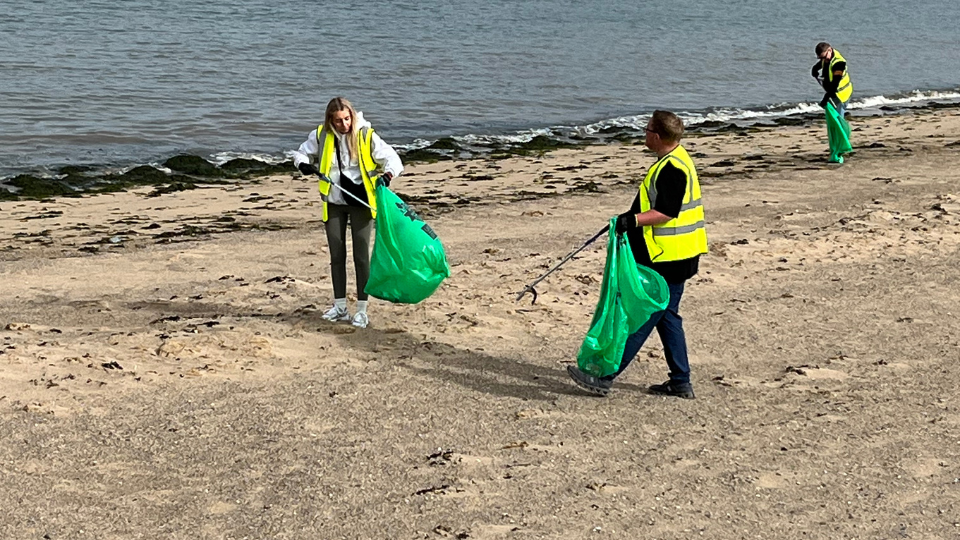 Three HW Facilities volunteers picking up litter on the beach, using grabber sticks and large green bin bags.